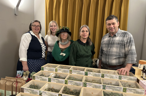 Skupina petih ljudi stoji za mizo na sejmu. Na mizi so razstavljene številne lesene škatle s posušenimi zelišči in čaji.