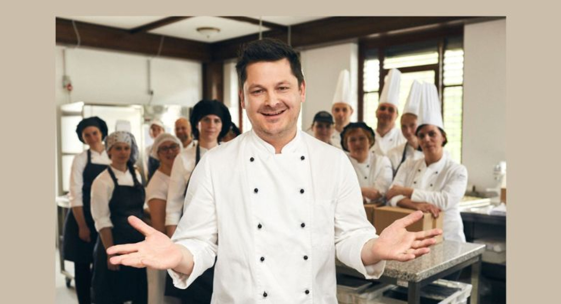 A man in a white chef's uniform, his team in the background with their arms spread wide.