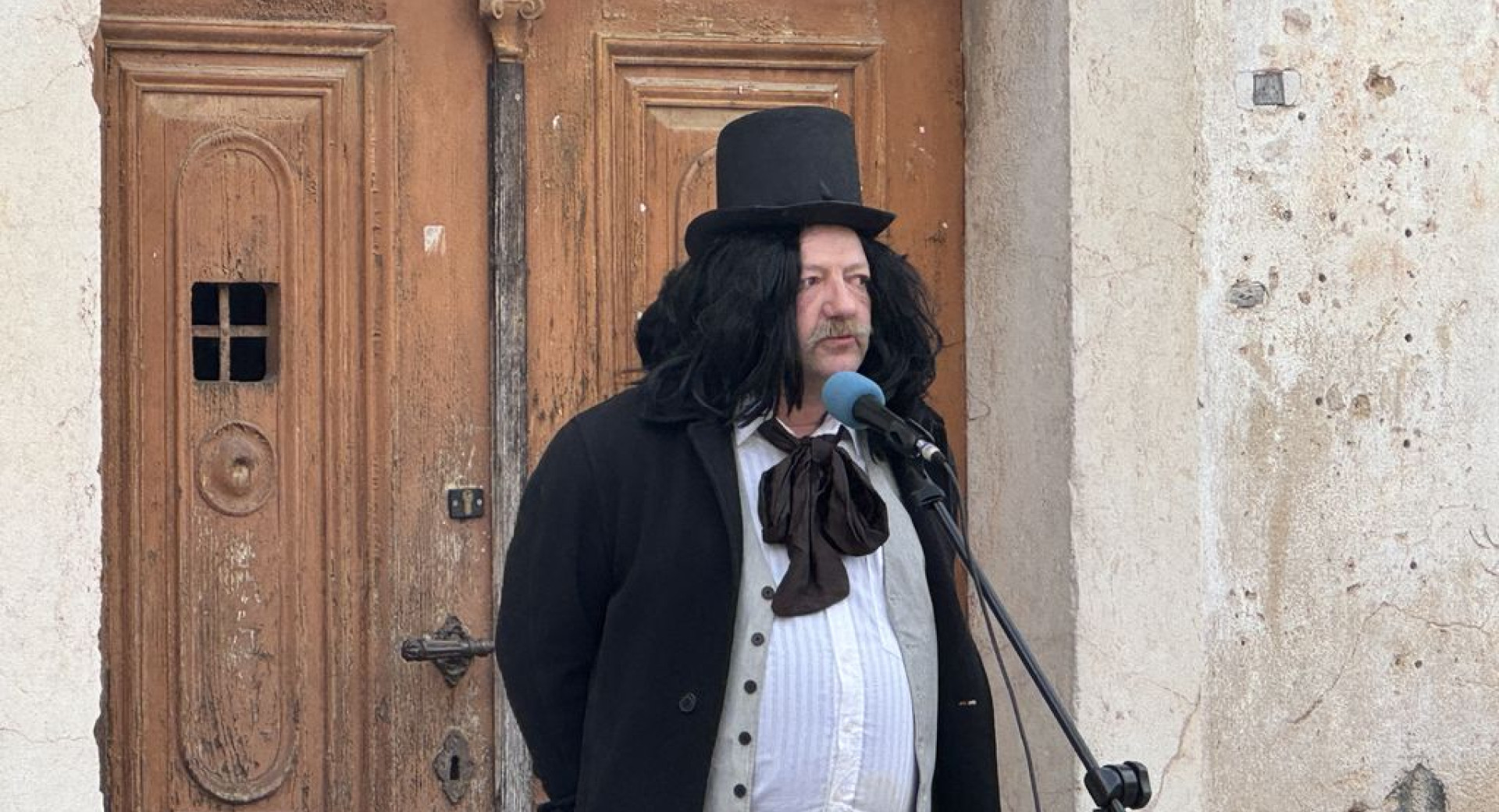 Moški v kostumu s cilindrom in v črnem suknjiču pred mikrofonom.