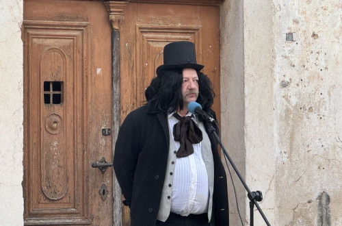 Moški v kostumu s cilindrom in v črnem suknjiču pred mikrofonom.