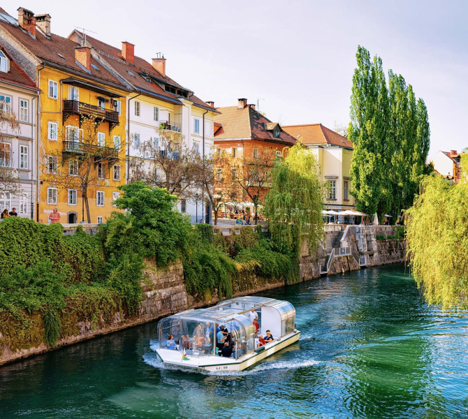Ladjica s turisti, ki pluje po reki, na levi strani nabrezje z zelenjem in mescanskimi hisami.