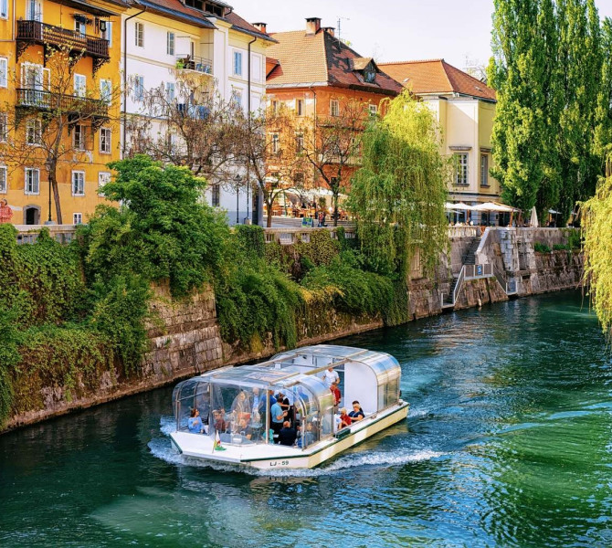 Ladjica s turisti, ki pluje po reki, na levi strani nabrezje z zelenjem in mescanskimi hisami.