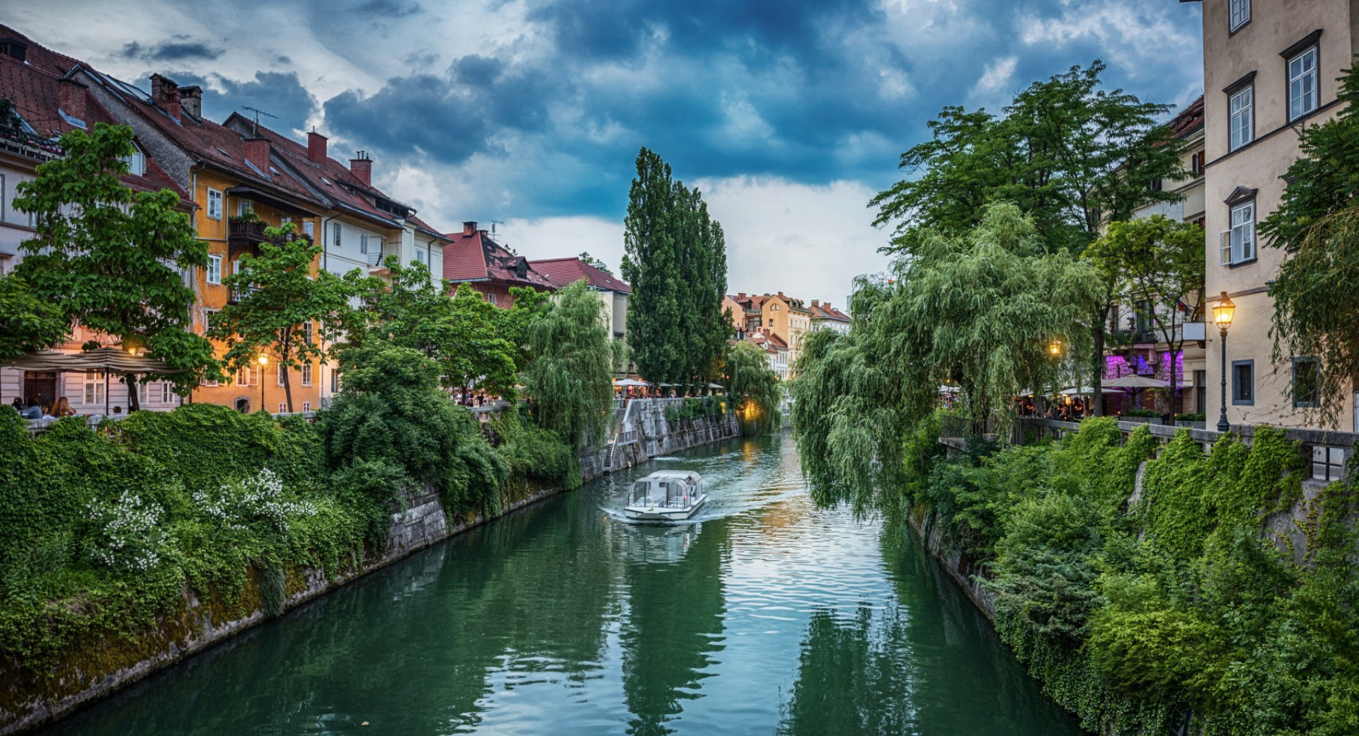 A boat sailing down the river, flanked by green embankments and bourgeois houses on both sides.