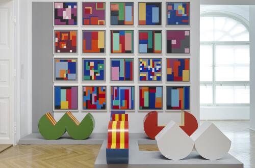 V galerijskem prostoru je na steni razstavljena mreza barvitih geometrijskih slik, pred njo pa stoji vec abstraktnih tridimenzionalnih skulptur v zivih barvah.