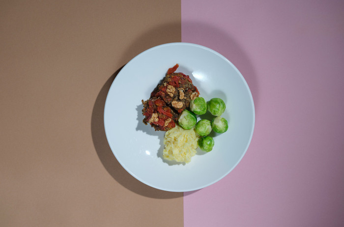 A white plate on a brown and pink base. On the plate, there is a neatly arranged roast with lentils and walnuts, alongside Brussels sprouts and mashed potatoes.