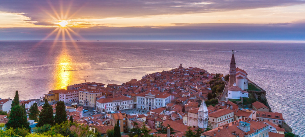 Panoramski pogled na slikoviti Piran ob sončnem zahodu, kjer sončni žarki obsijejo rdeče strehe mesta in Jadransko morje.