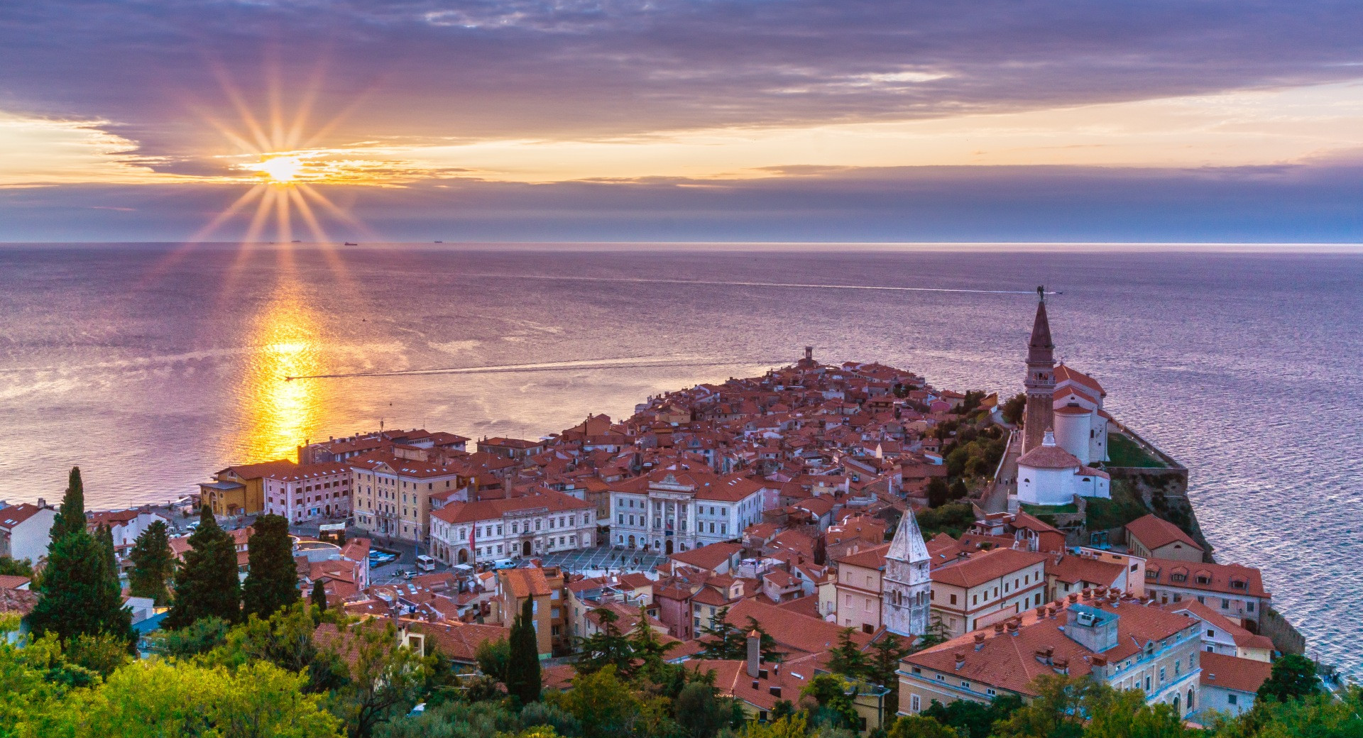 Panoramski pogled na slikoviti Piran ob sončnem zahodu, kjer sončni žarki obsijejo rdeče strehe mesta in Jadransko morje.