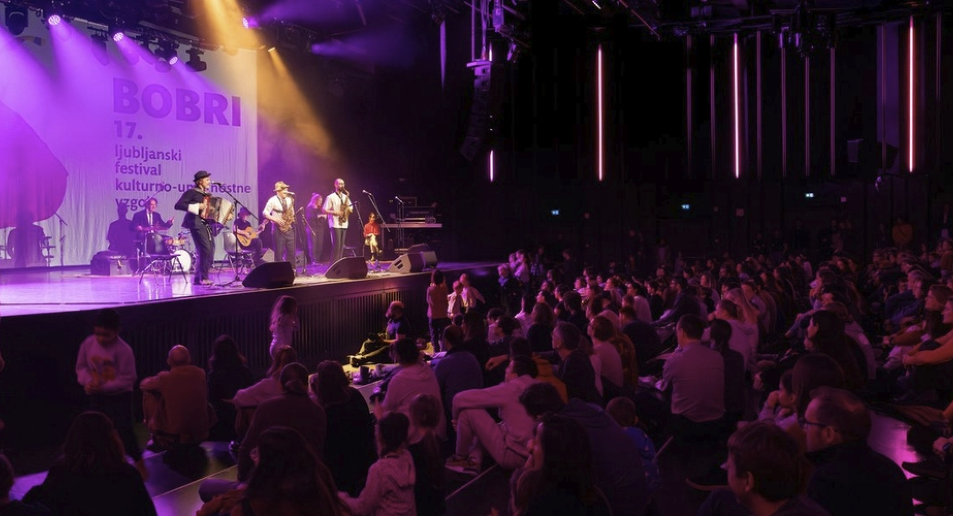 Koncert: glasbeniki na odru, pred njimi občinstvo.