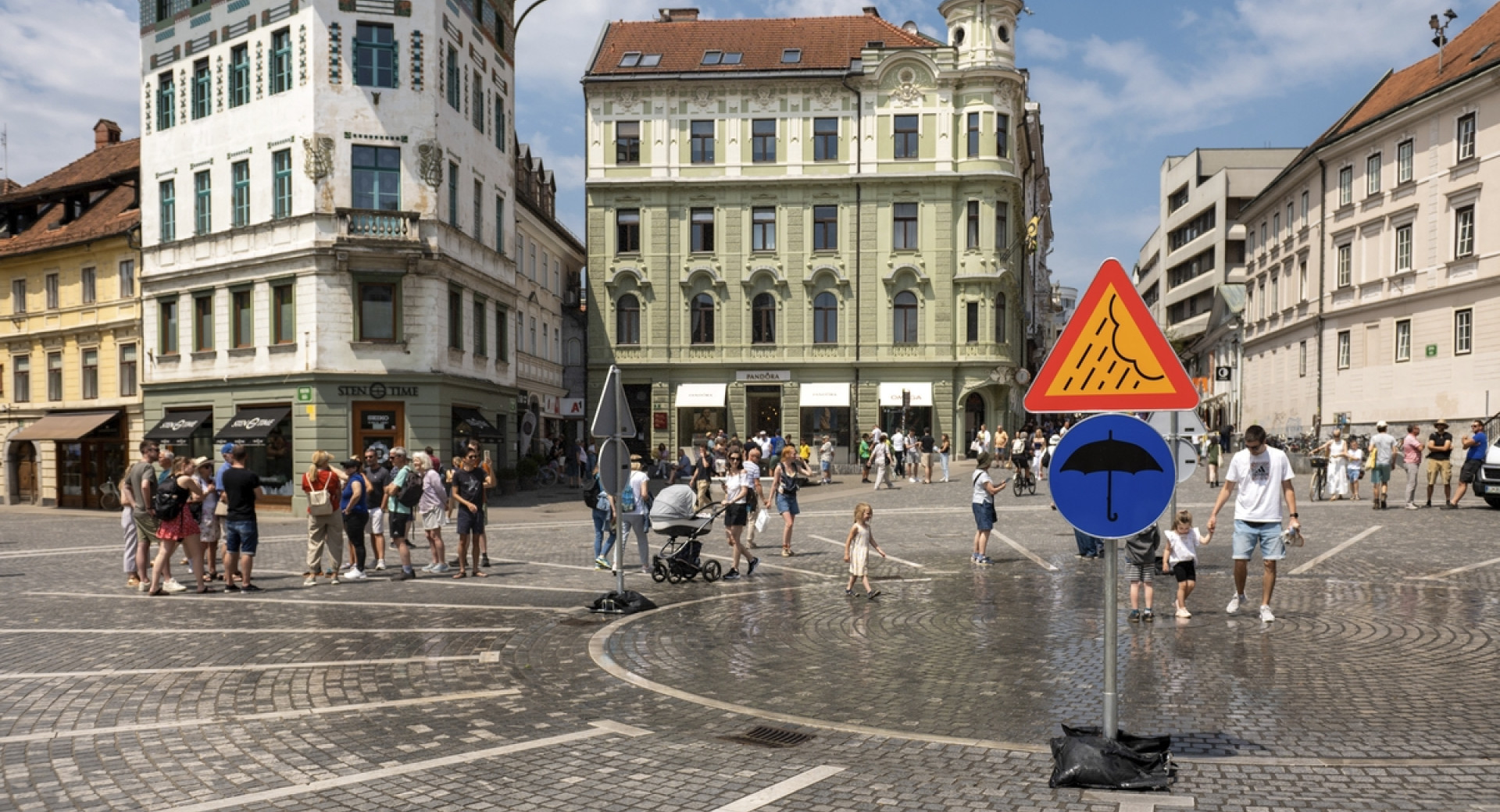 Trg sredi mesta, na njem ljudje in table z znakom za dežnik in dež.