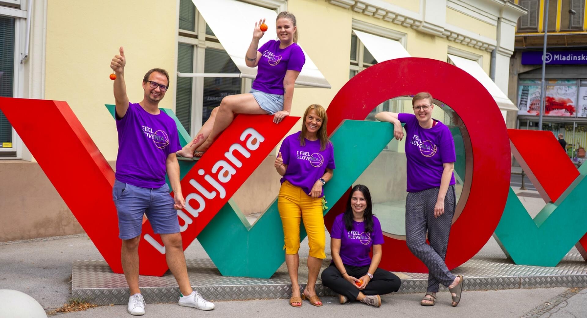 Skupina petih oseb v vijoličnih majicah "I Feel Slovenia" pozira ob velikem napisu Ljubljana, kažejo palce gor in držijo majhne košarkaške žogice.