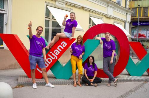 Skupina petih oseb v vijoličnih majicah "I Feel Slovenia" pozira ob velikem napisu Ljubljana, kažejo palce gor in držijo majhne košarkaške žogice.
