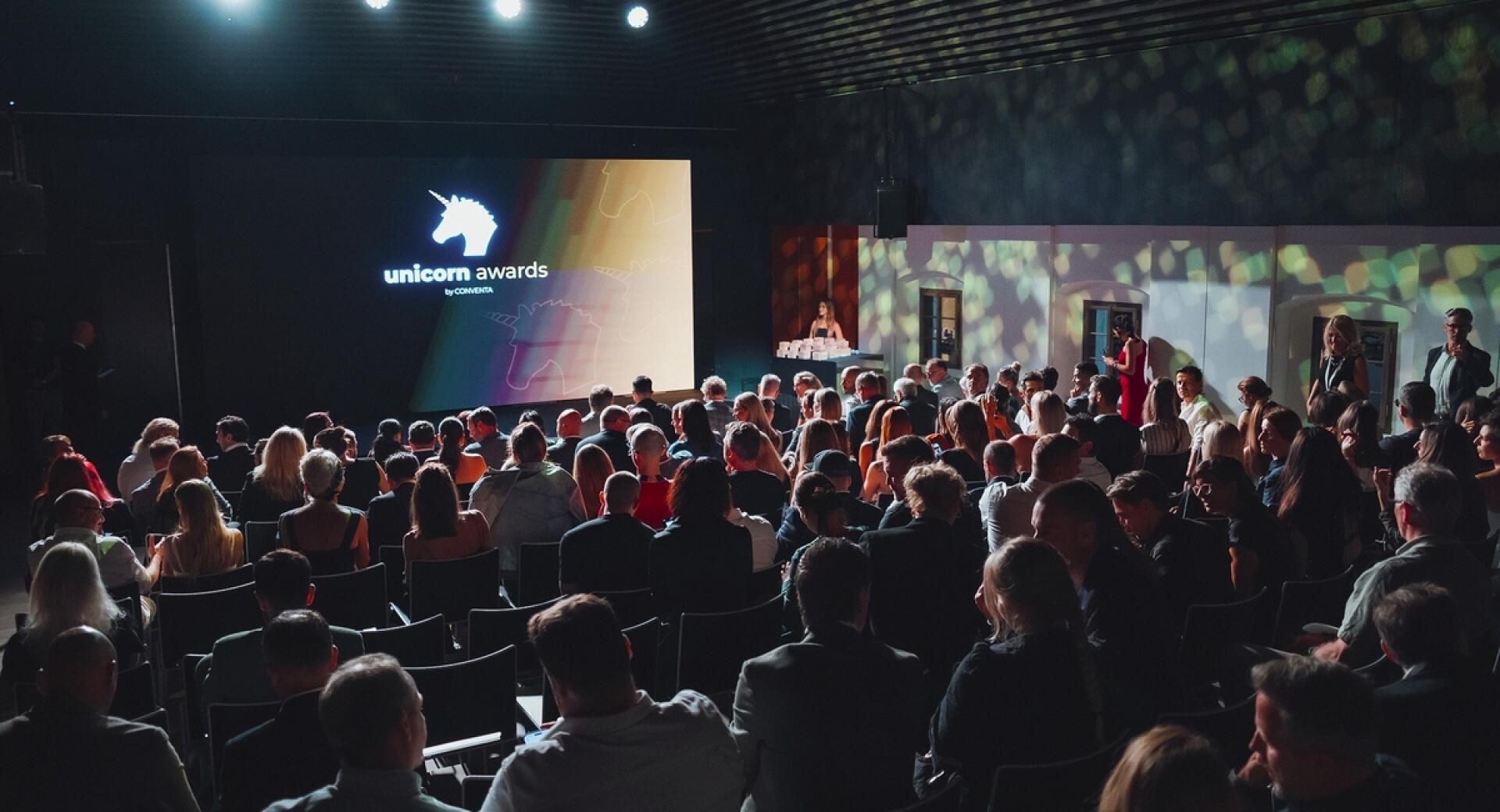 Občinstvo na dogodku Unicorn Awards v zatemnjeni dvorani. Na odru stoji govornik ob velikem zaslonu z logotipom dogodka.