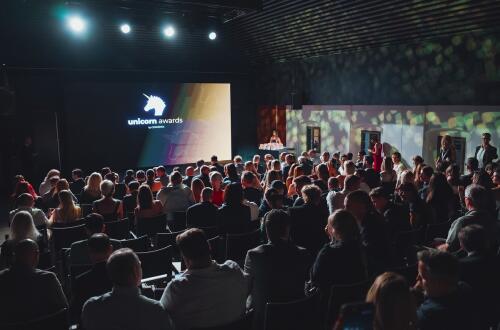 Občinstvo na dogodku Unicorn Awards v zatemnjeni dvorani. Na odru stoji govornik ob velikem zaslonu z logotipom dogodka.