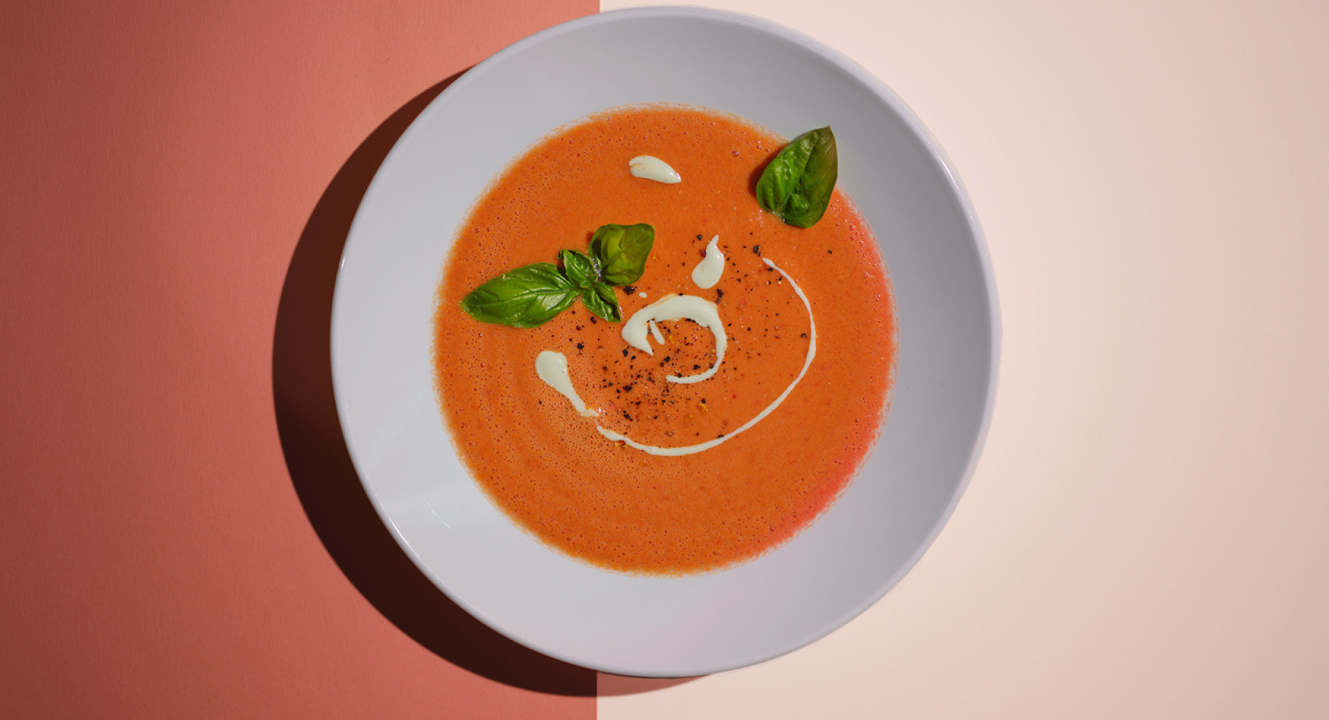 A white plate on a light and dark orange base. The plate is filled with tomato soup, with a cream spiral in the middle, adorned with basil leaves.