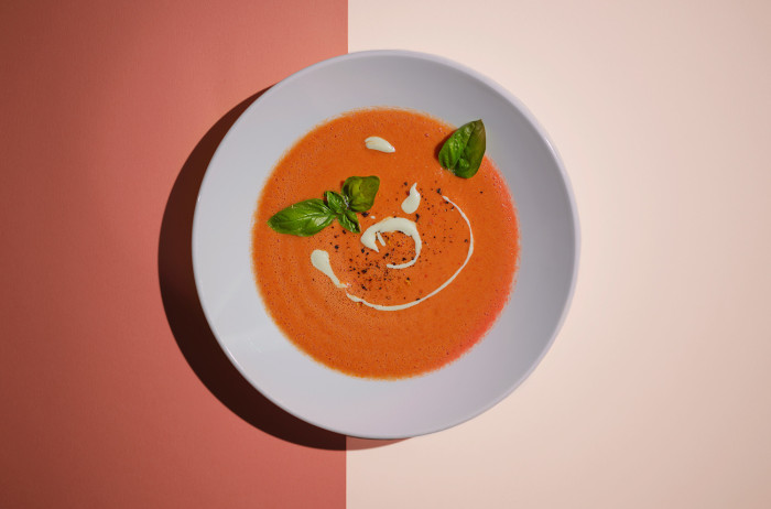 A white plate on a light and dark orange base. The plate is filled with tomato soup, with a cream spiral in the middle, adorned with basil leaves.