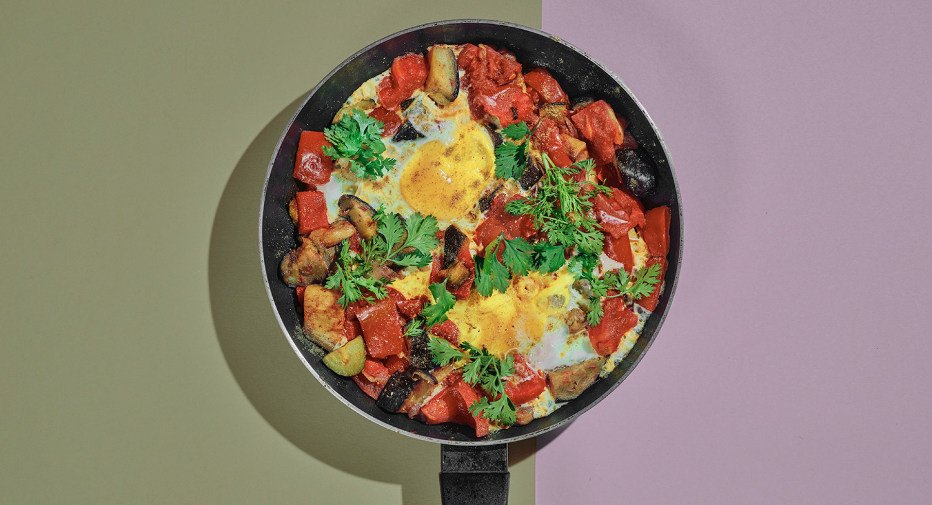 A black pan on an olive-green and violet base. In the pan, colorful sliced vegetables with eggs, sprinkled with parsley leaves.
