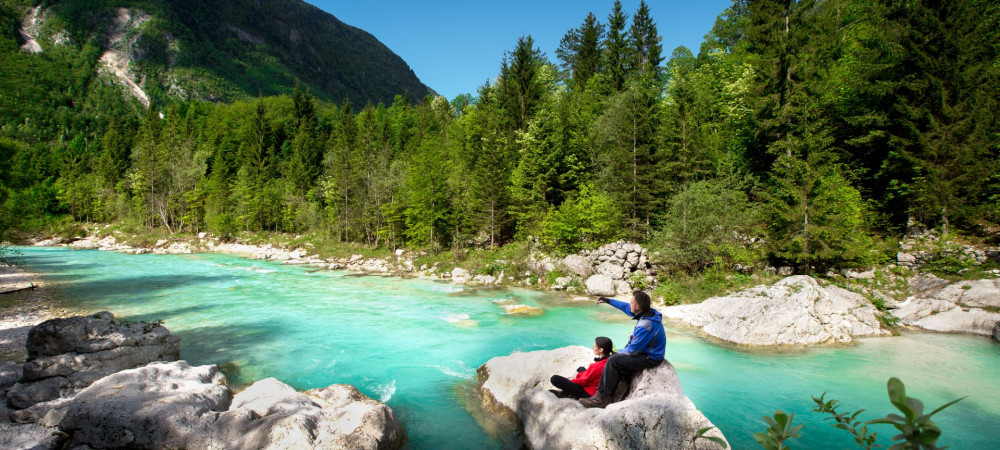 L'image montre un homme et une femme assis sur un rocher au milieu d'une rivière de montagne bleu turquoise, admirant le paysage.