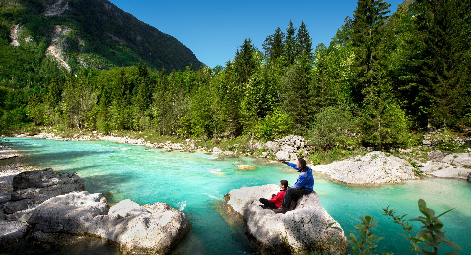 L'image montre un homme et une femme assis sur un rocher au milieu d'une rivière de montagne bleu turquoise, admirant le paysage.