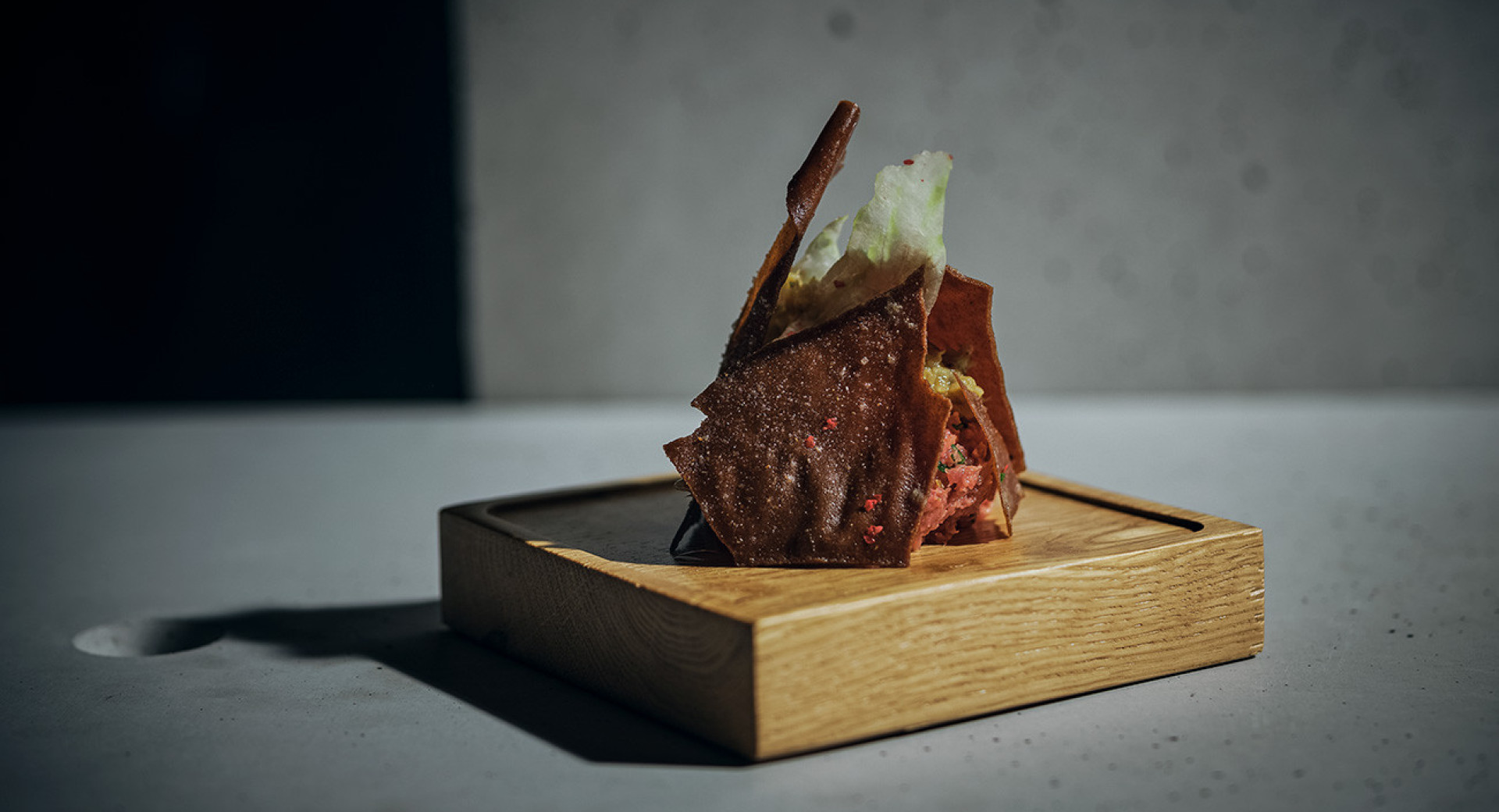 A modernly presented appetizer on a wooden board. Elevated crackers with tartare spread in between. White background with darkened edges in the photograph.