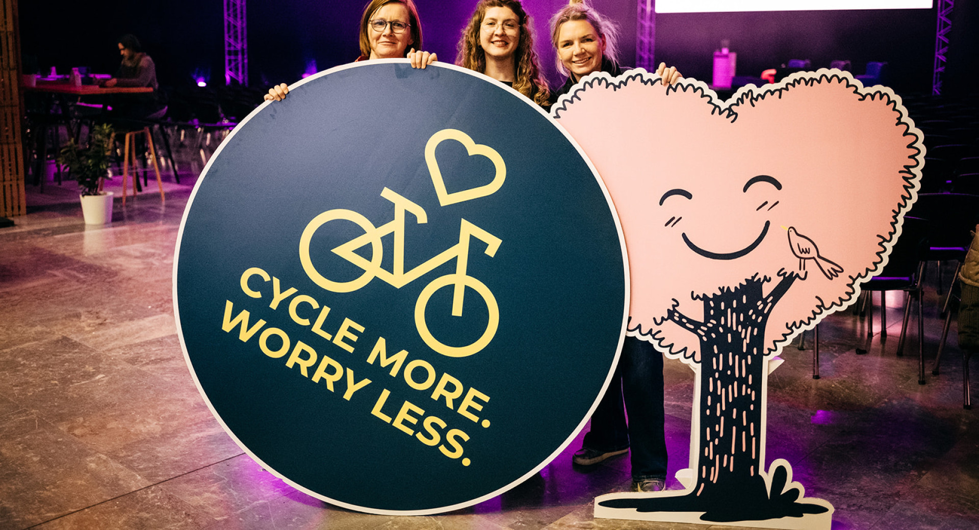 Tri osebe držijo v rokah okrogel pano z napisom: Cycle more. Worry less. (Več kolesarjenja. Manj skrbi.)