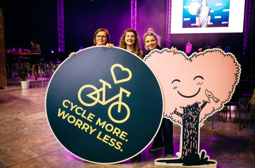 Tri osebe držijo v rokah okrogel pano z napisom: Cycle more. Worry less. (Več kolesarjenja. Manj skrbi.)