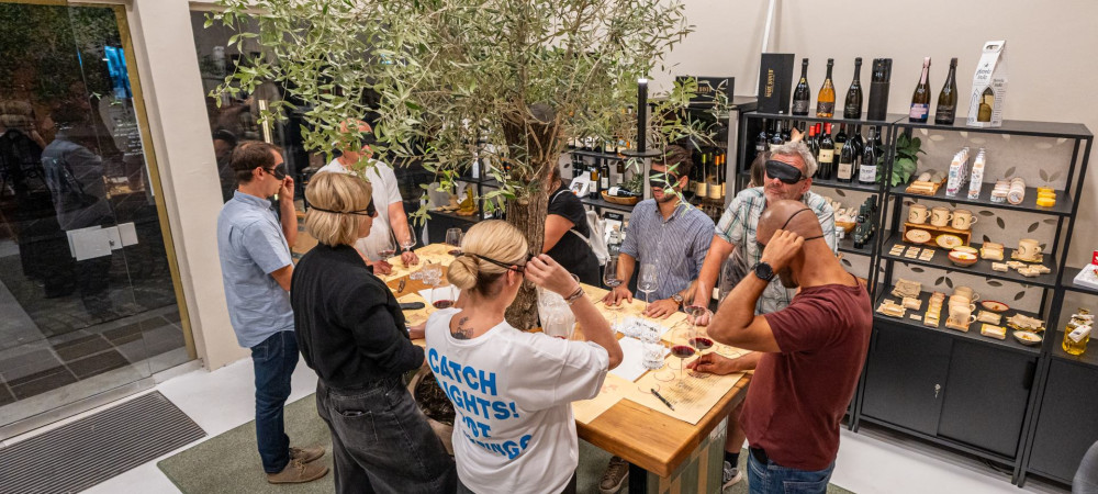 Un gruppo di persone è in piedi attorno a un tavolo, attraverso il quale cresce un albero, e stanno degustando diversi vini con le bende sugli occhi.