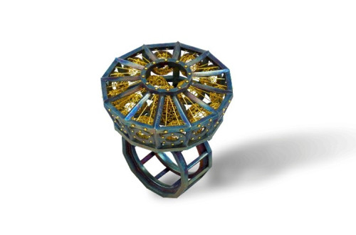 Umetniški osmerokoten prstan z modrikasto kovinsko kletko, v kateri so prepletene fine zlate niti in krogle, postavljen na dvojno geometrijsko ogrodje.
