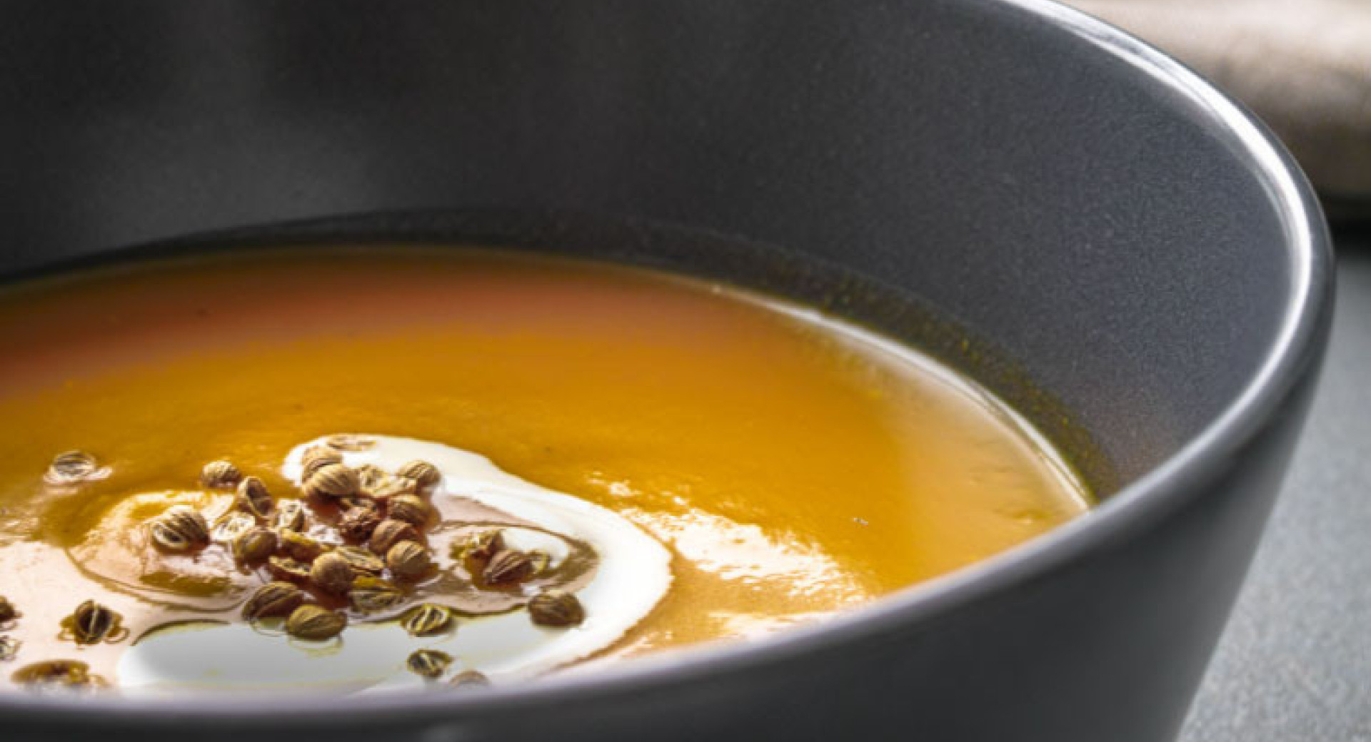 Pumpkin soup in a large black bowl, with a black base. In the orange soup, there's a white swirl of cream, garnished with pumpkin seeds.