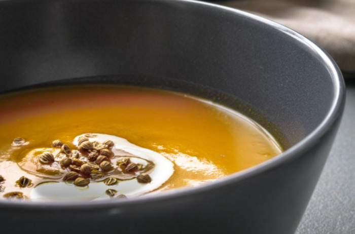 Pumpkin soup in a large black bowl, with a black base. In the orange soup, there's a white swirl of cream, garnished with pumpkin seeds.
