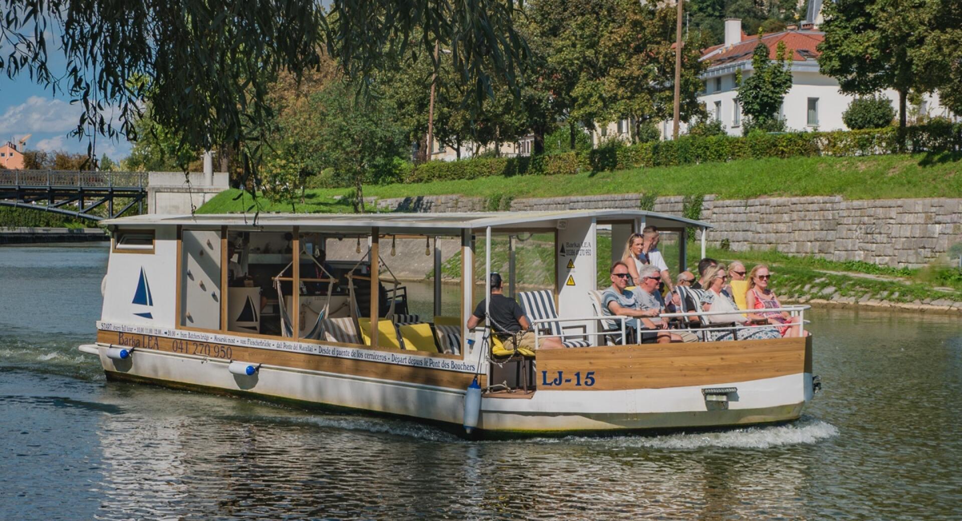 The tourist boat Barka Lea carrying passengers cruises along the Ljubljanica River by a green embankment, with Ljubljana Castle rising on the hill in the background.