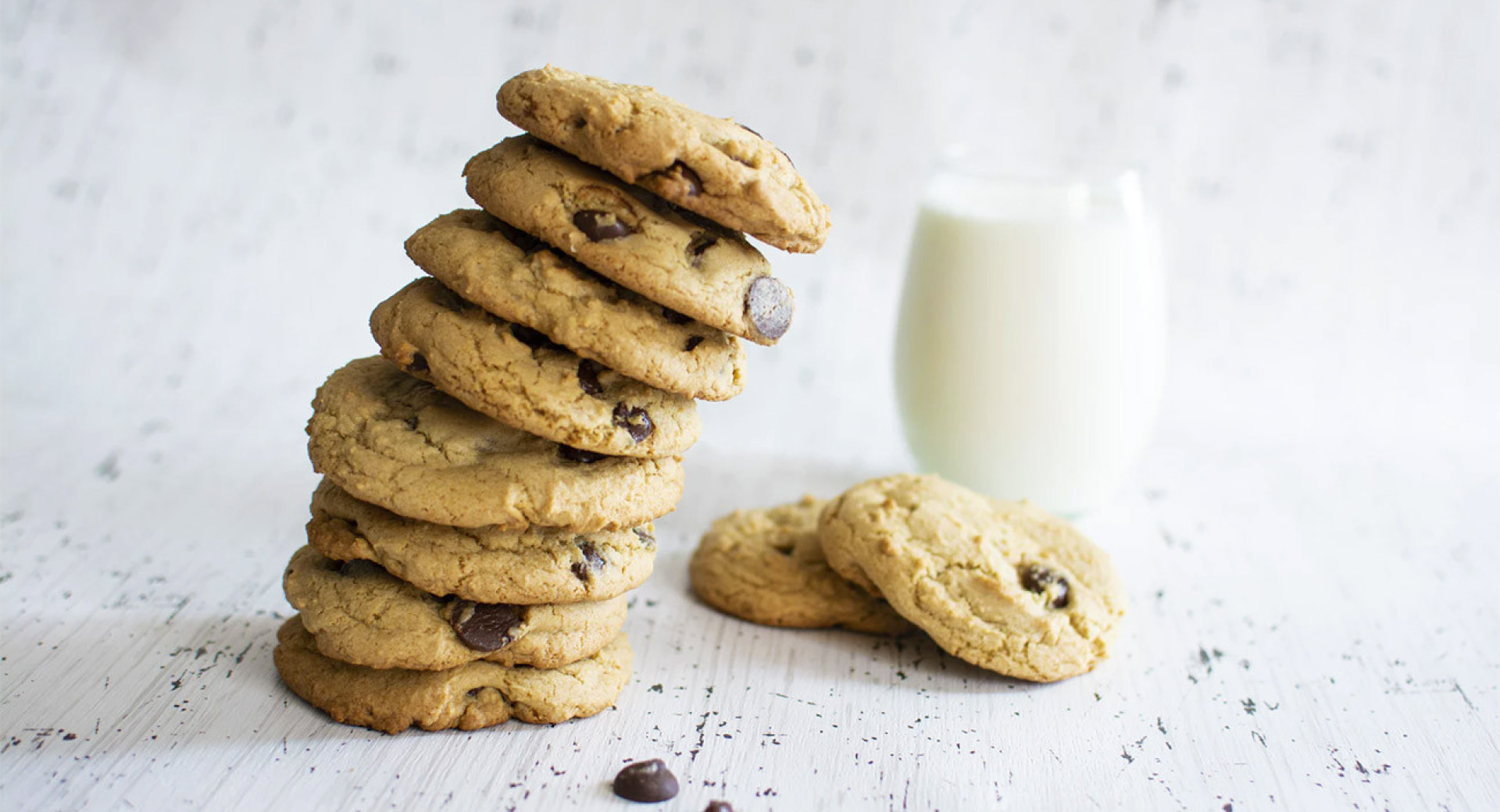 Cookies with chocolate chunks stacked into a tower. Besides two standalone cookies, there's also a glass of milk. The background is white.