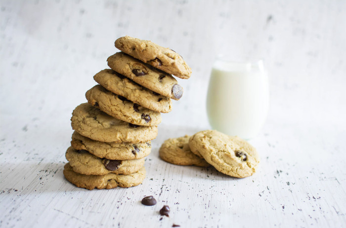 Cookies with chocolate chunks stacked into a tower. Besides two standalone cookies, there's also a glass of milk. The background is white.