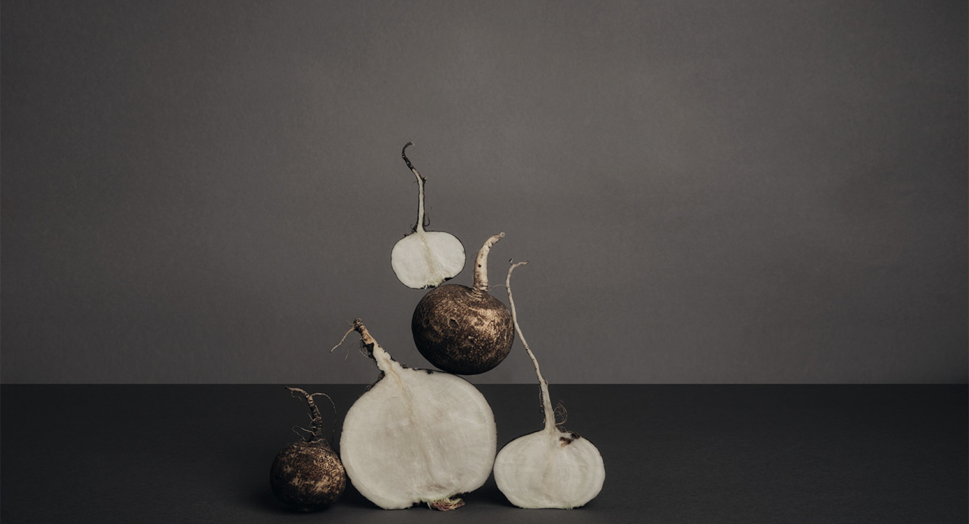 A whole and halved radish arranged in a pyramid shape. Black background, gray backdrop.