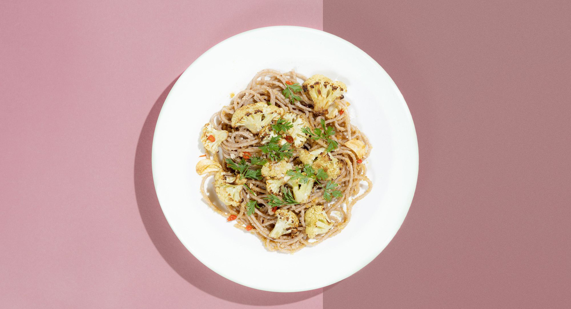 A white plate on a light and dark pink background. On the plate, there's a mound of spaghetti decorated with cauliflower florets and parsley.