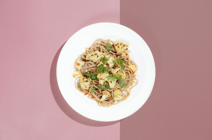A white plate on a light and dark pink background. On the plate, there's a mound of spaghetti decorated with cauliflower florets and parsley.