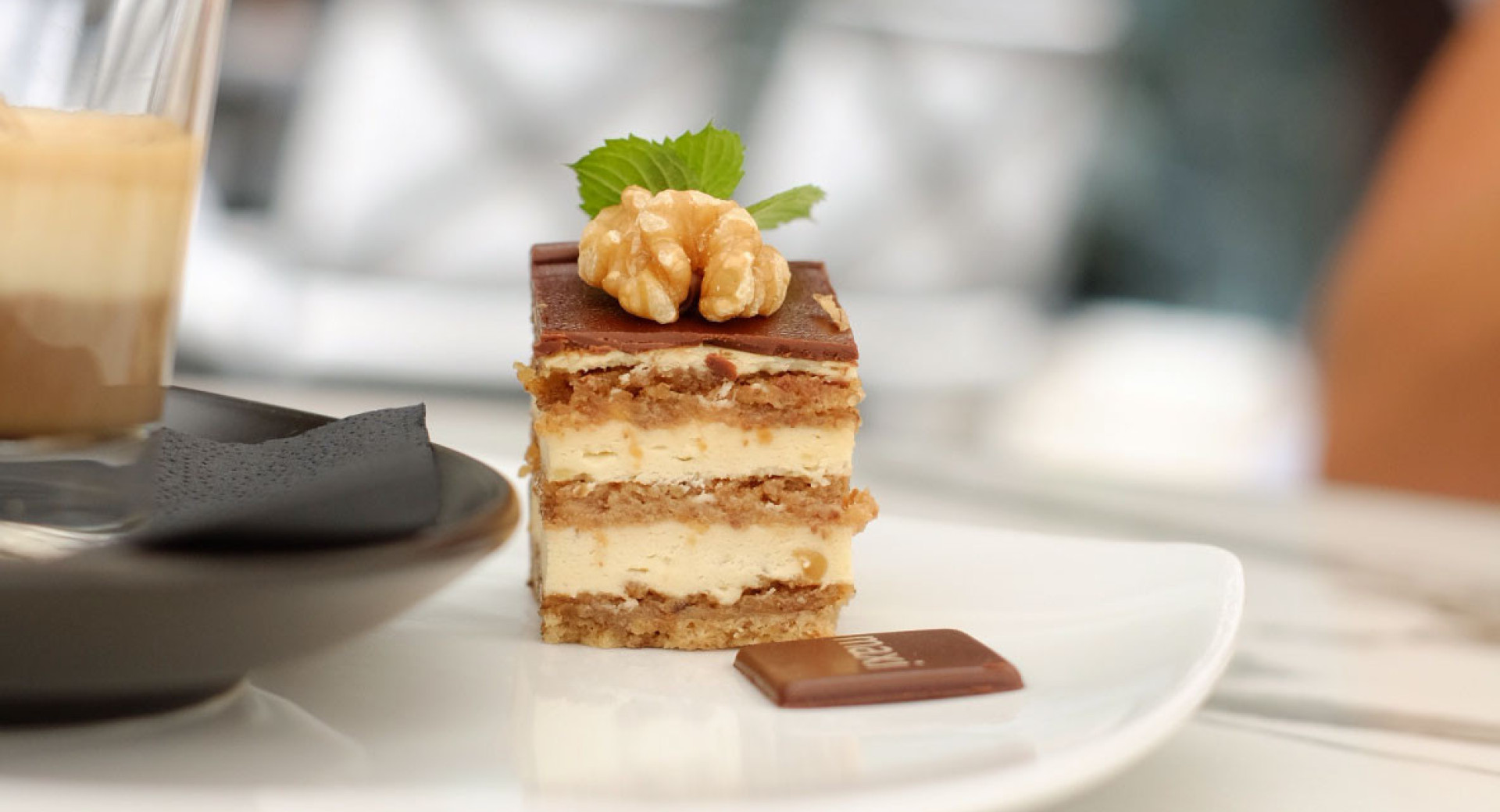 A cube of pastry on a white plate next to a cup of coffee. The pastry consists of sponge cake and cream, decorated with a walnut kernel and a mint leaf. In the background, out of focus, are restaurant chairs.