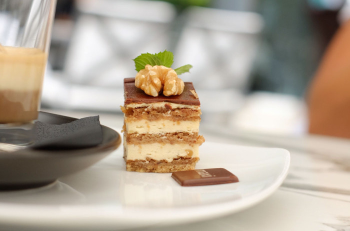 A cube of pastry on a white plate next to a cup of coffee. The pastry consists of sponge cake and cream, decorated with a walnut kernel and a mint leaf. In the background, out of focus, are restaurant chairs.