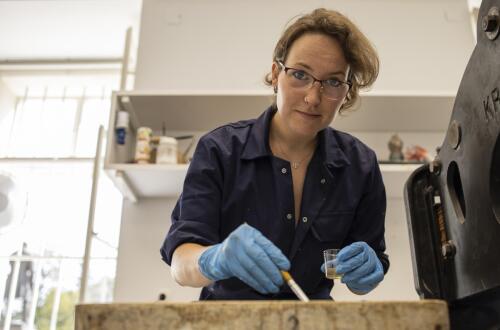 A person wearing gloves in a studio demonstrates the application of a liquid onto a linocut plate.