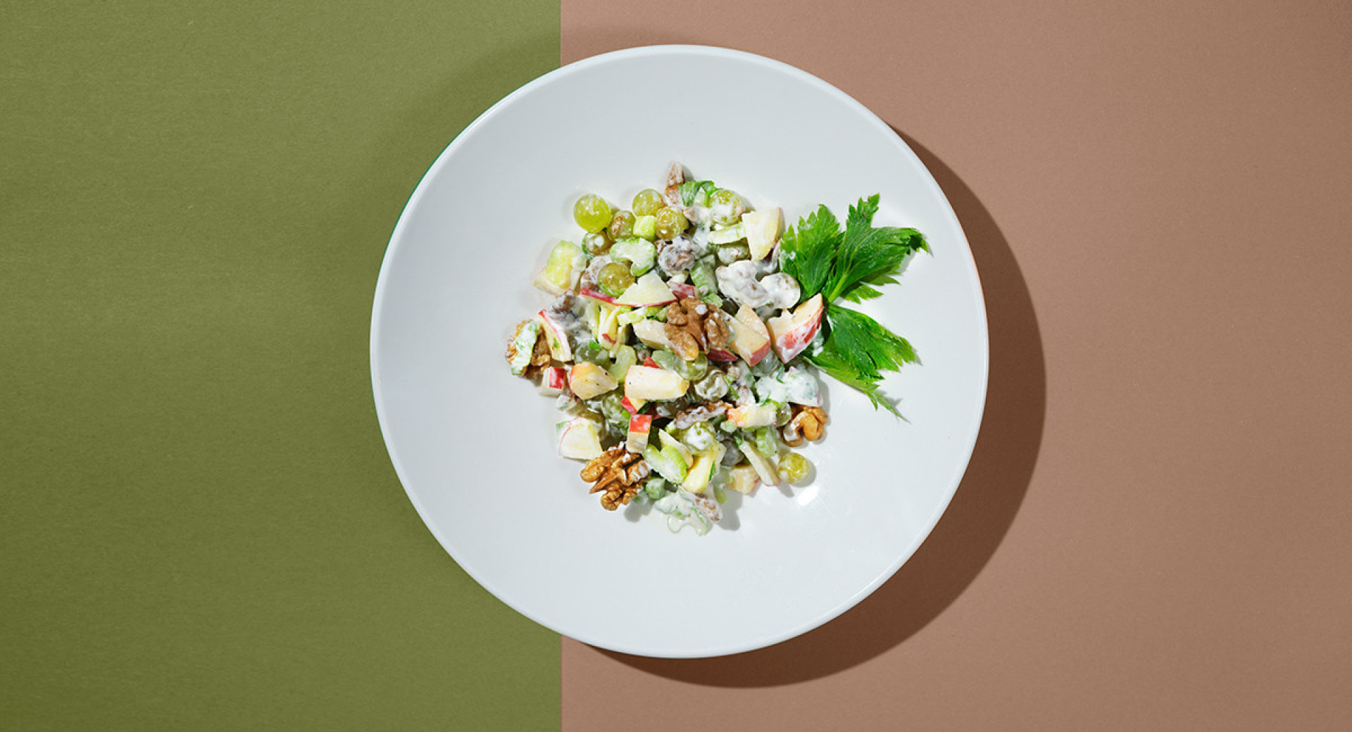  A white plate on an olive green and pink base. On the plate, a nicely presented salad with dressing, decorated with walnuts and parsley leaves.