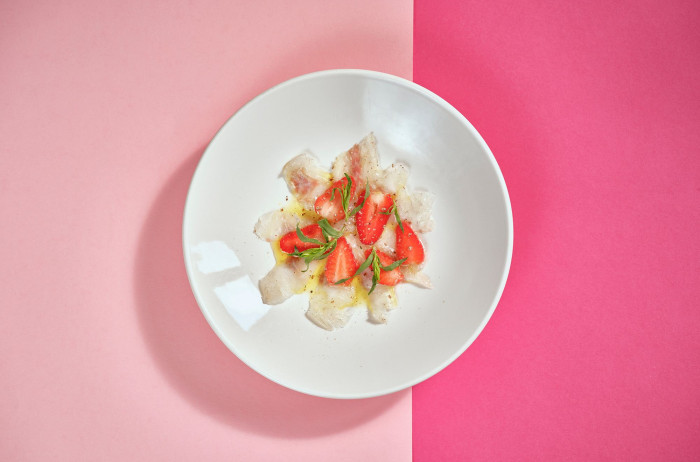 A white plate on a light and dark pink base. On the plate, neatly arranged fish, decorated with strawberries and greens.