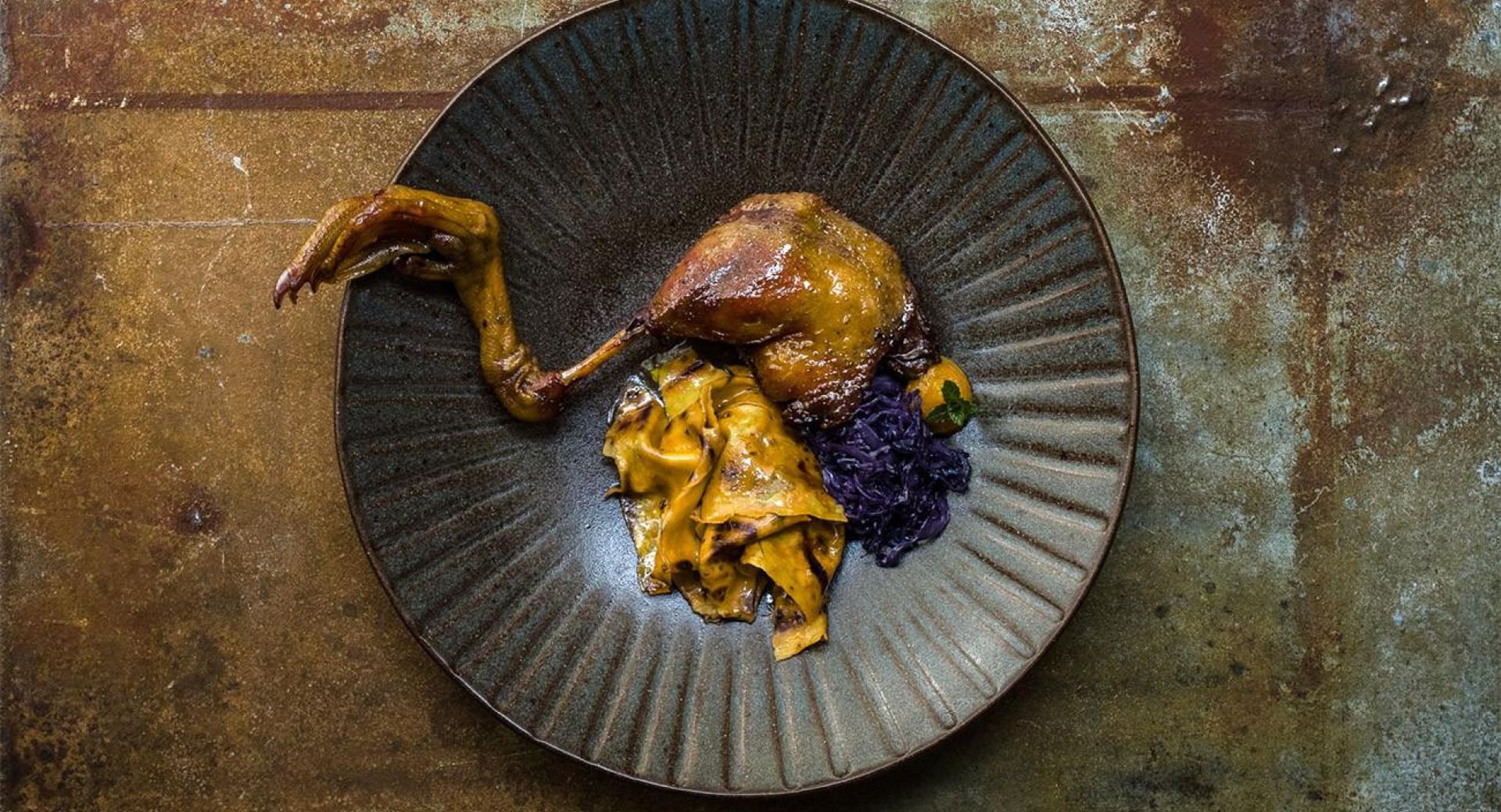 Duck leg placed over a dark plate. Next to it, there are pasta squares (known as mlinci) and red cabbage. The plate is on a brown, rustic base.