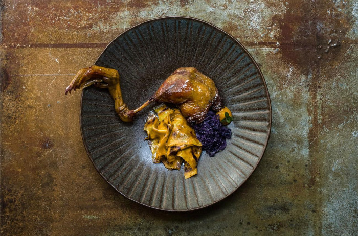 Duck leg placed over a dark plate. Next to it, there are pasta squares (known as mlinci) and red cabbage. The plate is on a brown, rustic base.