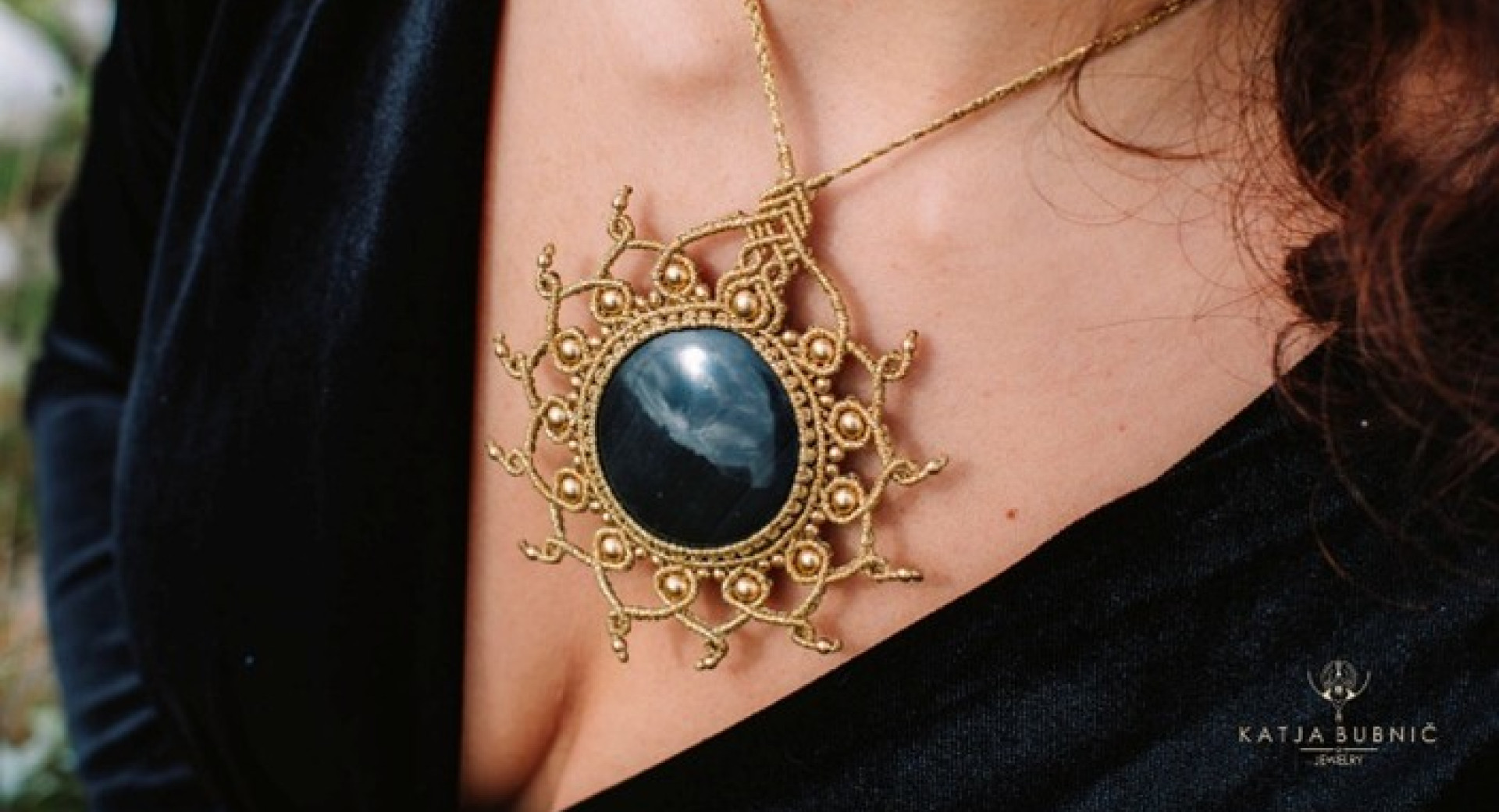 A big necklace with a large black stone and a golden macramé braided sun-shaped frame, worn by a person in a black dress.