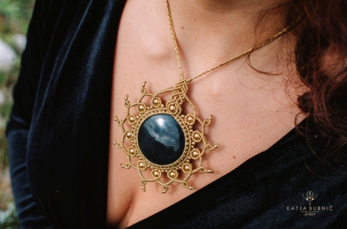 A big necklace with a large black stone and a golden macramé braided sun-shaped frame, worn by a person in a black dress.