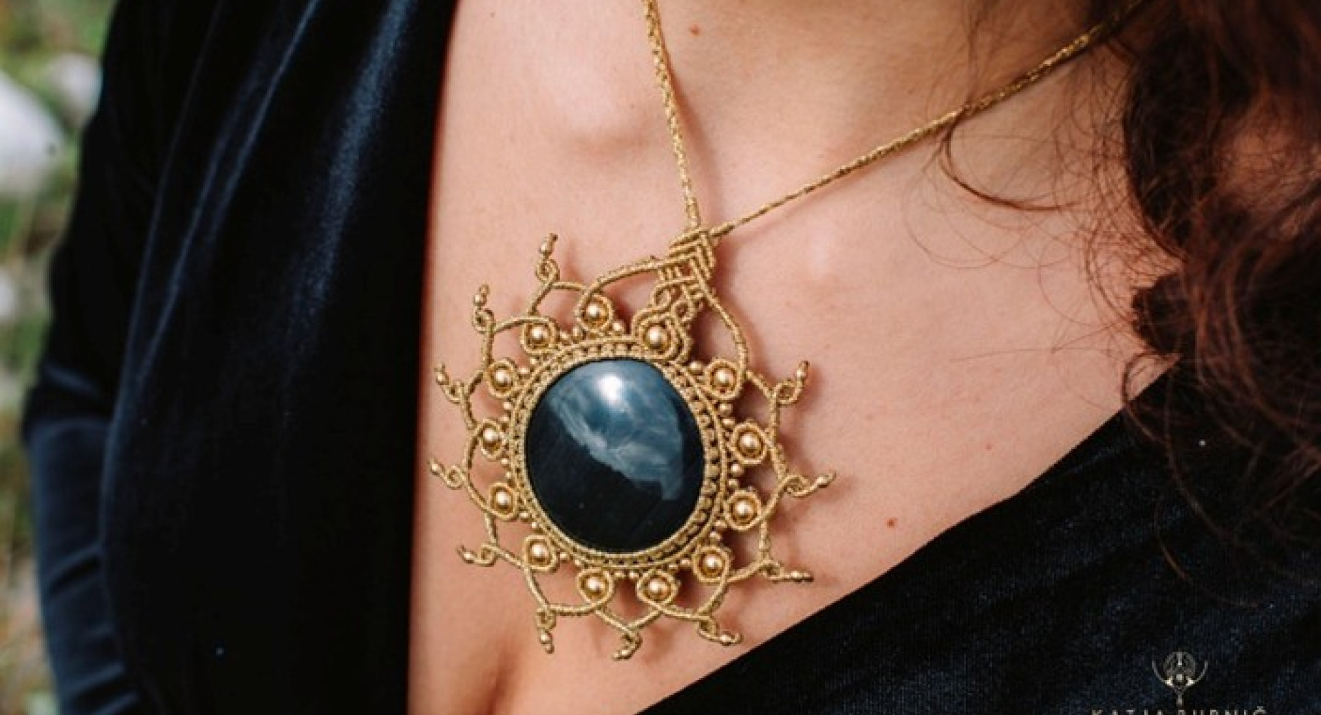 Masivna ogrlica z velikim črnim kamnom v sredini, obdanim z bogato pletenim zlatim okvirom v obliki sonca z ukrivljenimi žarki, ki jo nosi oseba v črni žametni obleki.