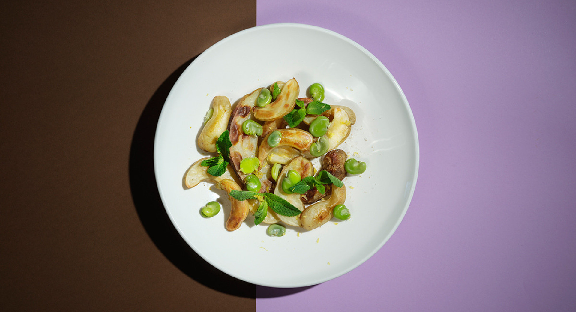 A white plate on a brown and violet base. On the plate, there's roasted potatoes sprinkled with green peas.