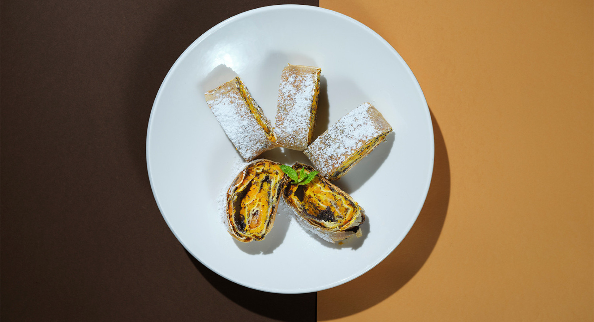 A white plate on a brown and orange base. On the plate, there are five pumpkin rolls neatly arranged, dusted with powdered sugar.