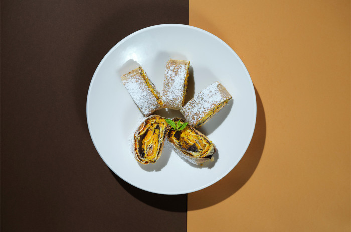A white plate on a brown and orange base. On the plate, there are five pumpkin rolls neatly arranged, dusted with powdered sugar.
