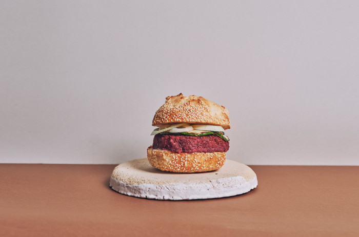 A stone plate on a brown table, white background. On the plate, there's a homemade burger presented, with a thick patty and vegetables between the slices of bread.