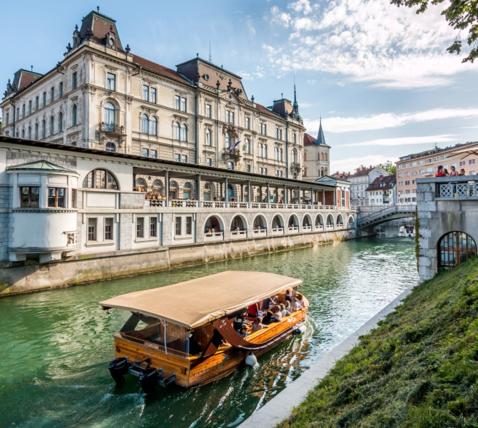 A wooden boat sailing on the river, with arcades and a large townhouse in the background.